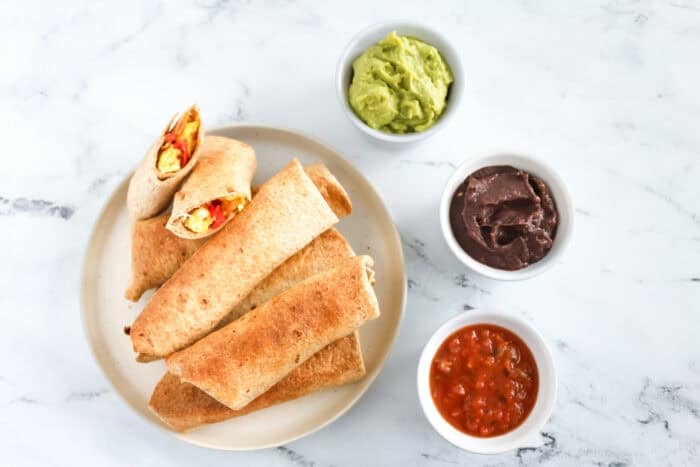 Plate of baked breakfast taquitos filled with scrambled eggs and peppers, served with ¡Yo Quiero! Grab & Go Guacamole, Black Bean Dip, and Salsa.
