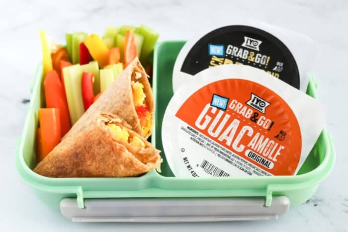 Lunchbox with veggie egg wrap, colorful bell pepper and celery sticks, and two ¡Yo Quiero! Grab & Go dips—guacamole and bean dip.