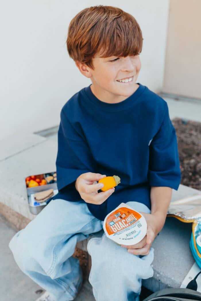 Smiling boy sitting outdoors holding a yellow pepper dipped in guacamole, with a ¡Yo Quiero! Grab & Go Guacamole cup in hand.