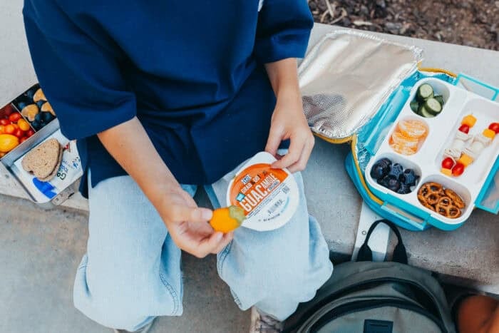 A boy sitting outdoors holding a yellow pepper dipped in guacamole, with a ¡Yo Quiero! Grab & Go Guacamole cup in hand.