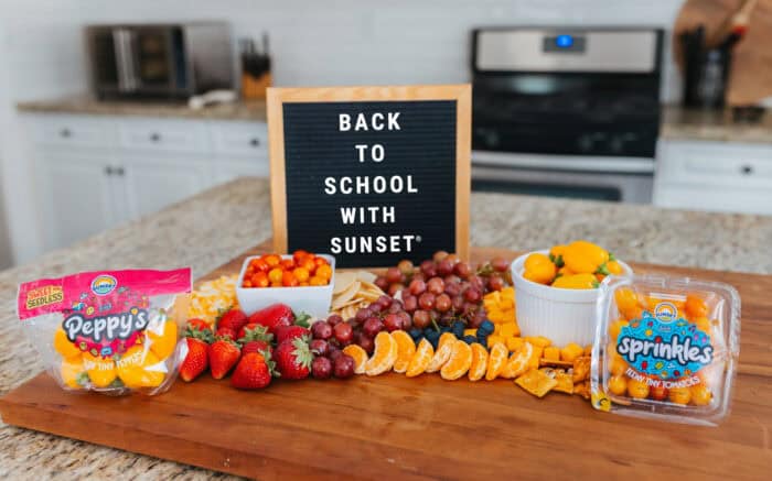 Back-to-school snack board on a kitchen island featuring Sunset® Sprinkles tomatoes, Peppy’s teeny tiny peppers, fruit, cheese cubes, and a letterboard sign reading “Back to School with SUNSET®.”