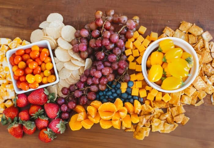 Overhead view of a colorful snack board with Sunset® Sprinkles tomatoes, yellow grape tomatoes, strawberries, grapes, blueberries, cheese cubes, mandarin slices, crackers, and tortilla chips on a wooden board.