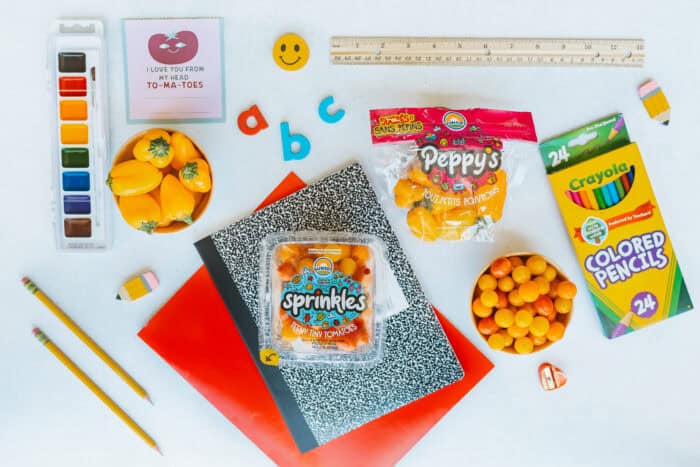 Flat lay of back-to-school supplies including Sunset® Sprinkles tomatoes, Peppy's teeny tiny peppers, colored pencils, notebooks, paint set, pencils, ruler, and alphabet magnets on a white background.